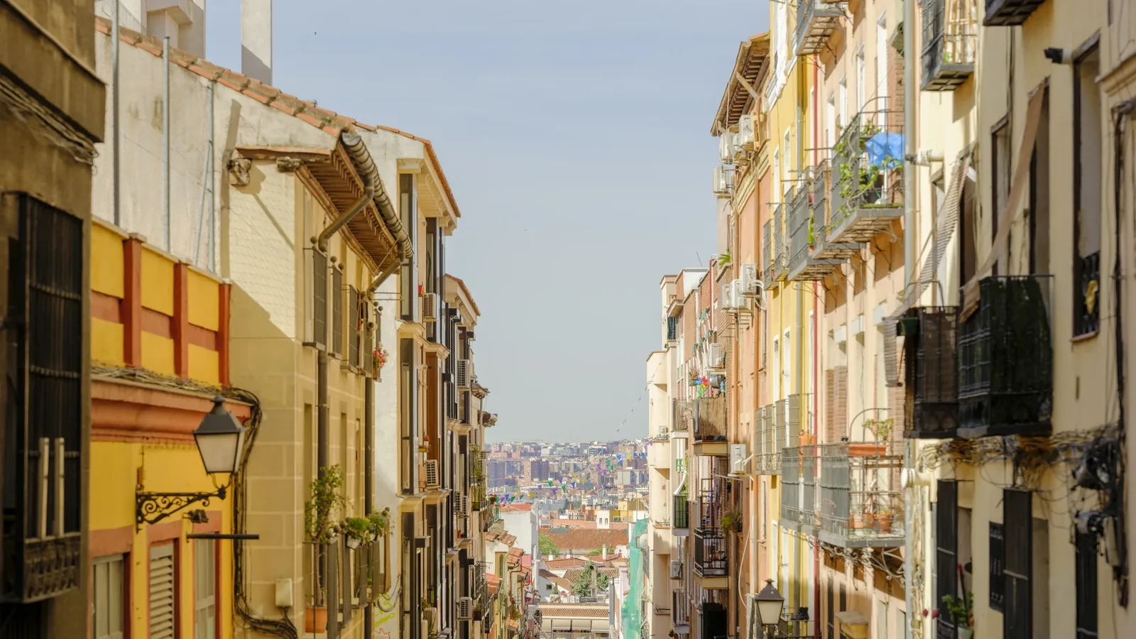 where to stay in madrid 4 Steep, narrow street in Lavapiés with pastel façades and iron balconies, the city skyline visible at the bottom of the hill